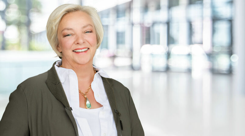 Portrait of a smiling businesswoman with short blonde hair wearing an olive green blazer and white shirt, in a bright office environment.